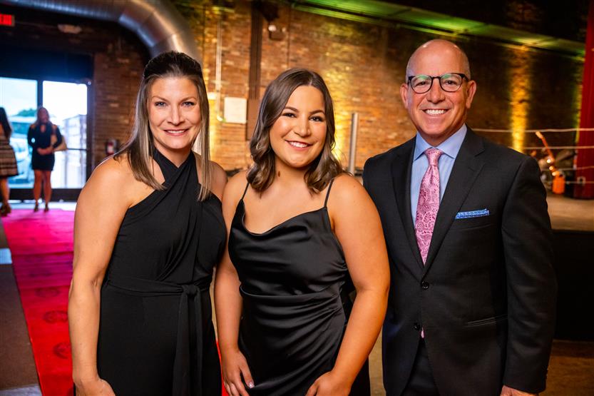 Two women wearing black dresses smile with a man wearing a black blazer and pink tie.